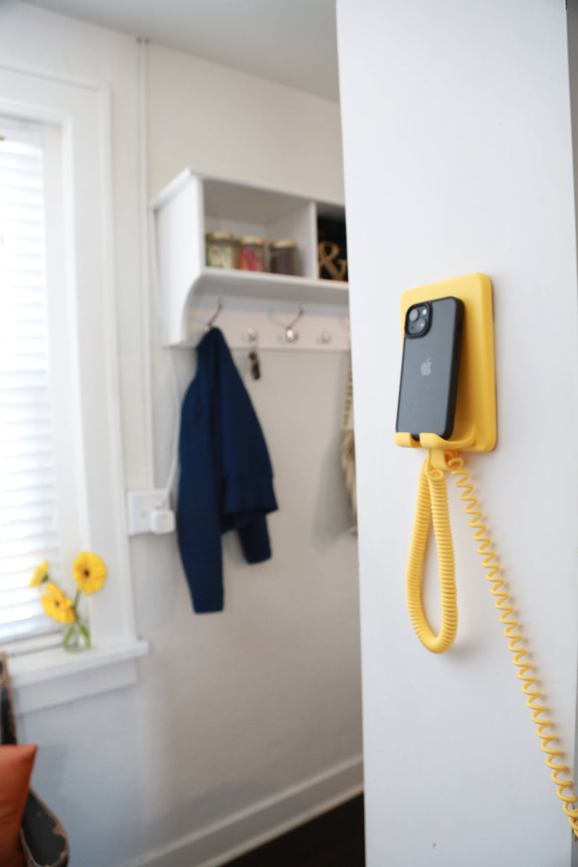 A yellow wall-mounted phone with a coiled cord and a black smartphone attached to its back, shown mounted on a white wall in a real interior setting with a coat rack, window, and jacket in the background.