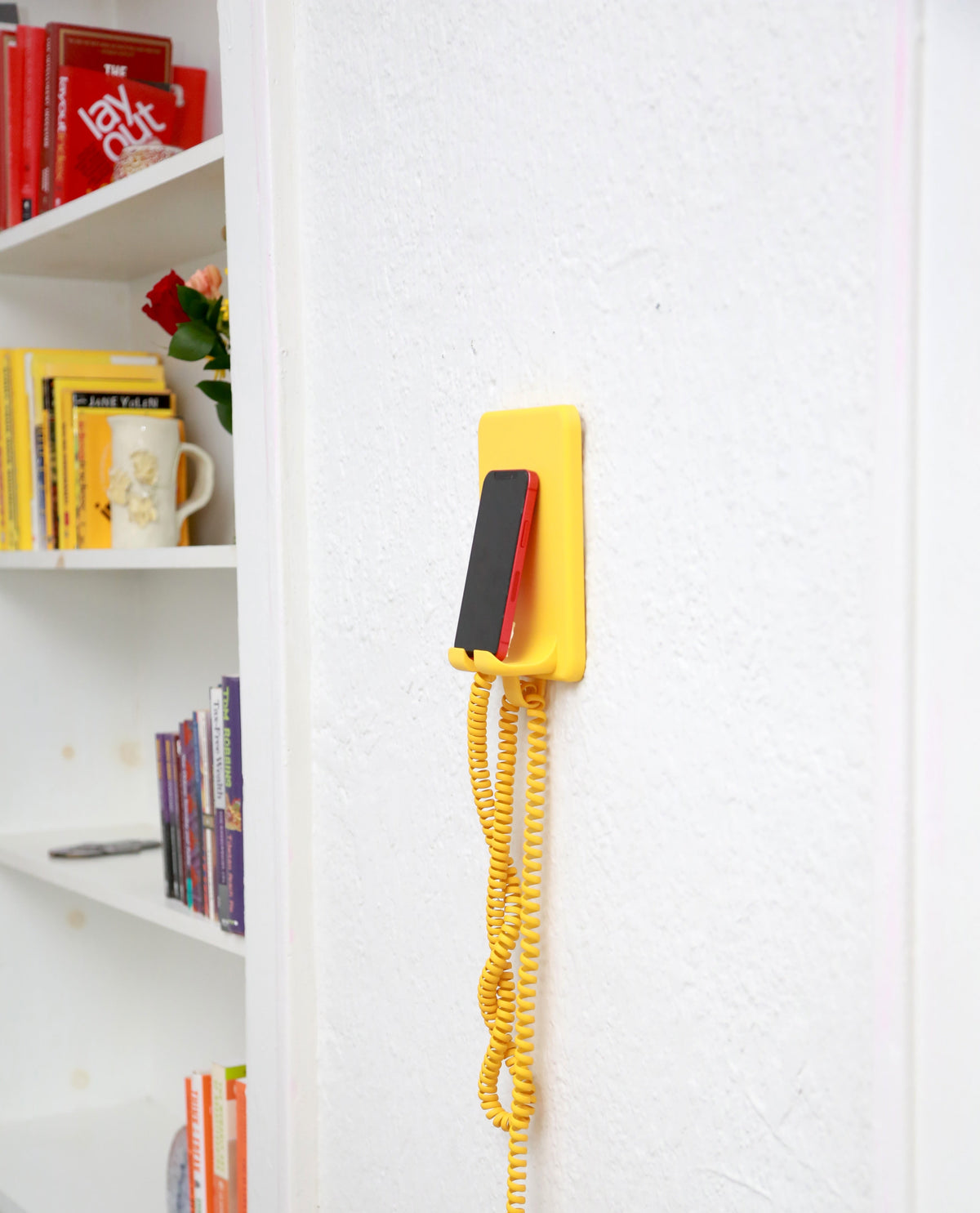 A retro-style wall-mounted telephone with a bright yellow base and coiled cord, holding a black and red phone, mounted on a white textured wall next to a white bookshelf.