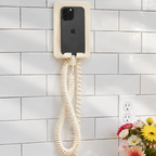 A wall-mounted cream-colored phone with a coiled cord and a black smartphone inserted into its cradle, mounted on a white subway tile wall above a wooden counter with flowers and a lemon.