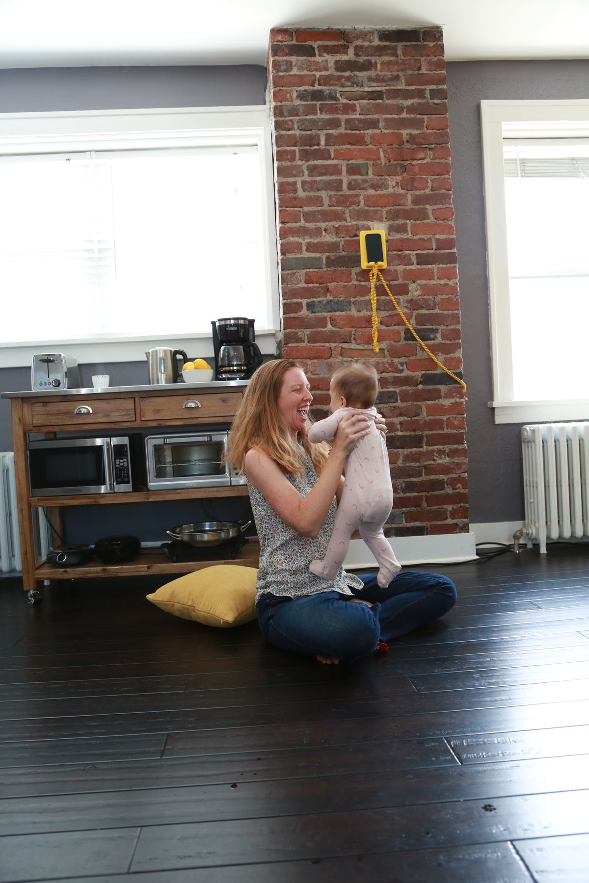 A woman sitting on a dark wood floor in a modern kitchen, holding a baby. The kitchen features a prominent red brick column, gray walls, and a wooden counter with a microwave and coffee maker.