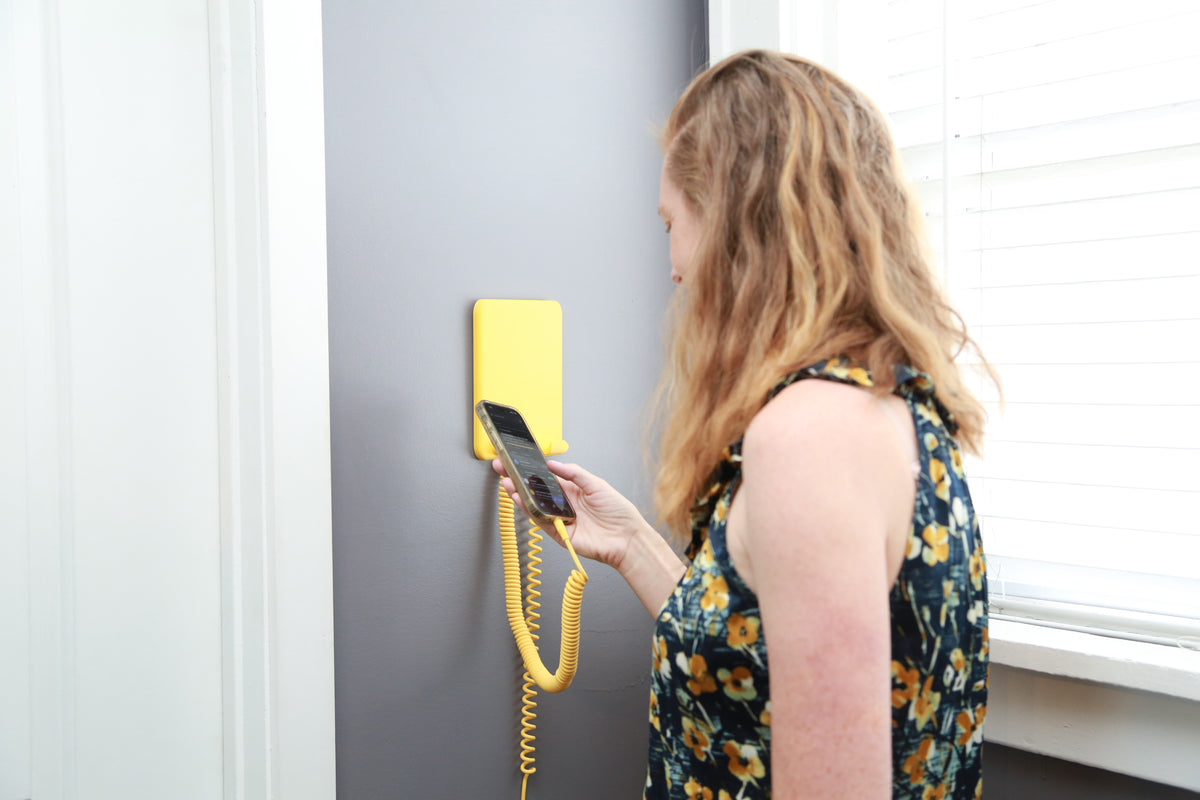A woman with long blonde hair stands in profile next to a window, holding a smartphone against a yellow wall-mounted telephone receiver with a coiled cord attached to a gray wall.